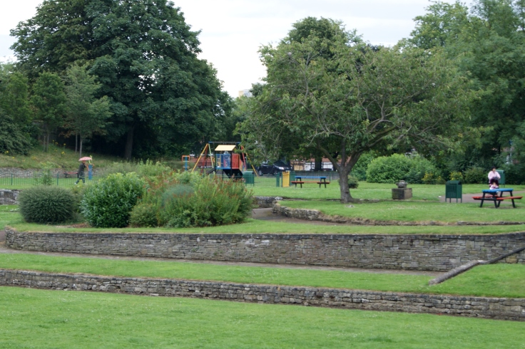 A playground with a man walking through it and people sat on a bench next to it.